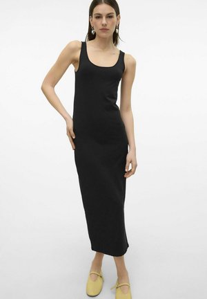 Robe midi noire sans manches avec une coupe ajustée, texture lisse et fentes latérales pour faciliter le mouvement, associée à des chaussures jaunes à enfiler.