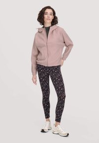 Giacca in pile rosa chiaro con zip e cappuccio, abbinata a leggins neri con stampa animale colorata e scarpe da ginnastica neutre.