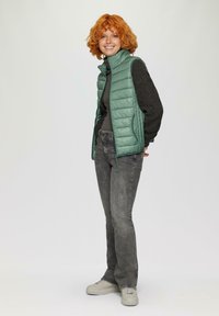 Hellgrüne Steppweste mit einem hohen Kragen, kombiniert mit einem schwarzen Langarmshirt und grauen Jeans, getragen mit hellgrauen Sneakers.