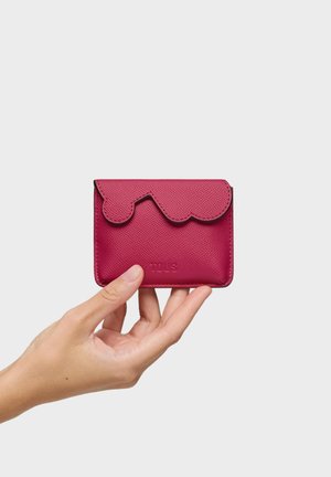 Dłoń trzymająca mały teksturowany czerwony skórzany portfel z falistą klapką i wytłoczonym nazwiskiem marki na jednolitym tle.