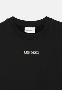 Sweat-shirt noir avec un col côtelé, arborant "LES DEUX" brodé en doré sur le devant. Tissu doux et design épuré.