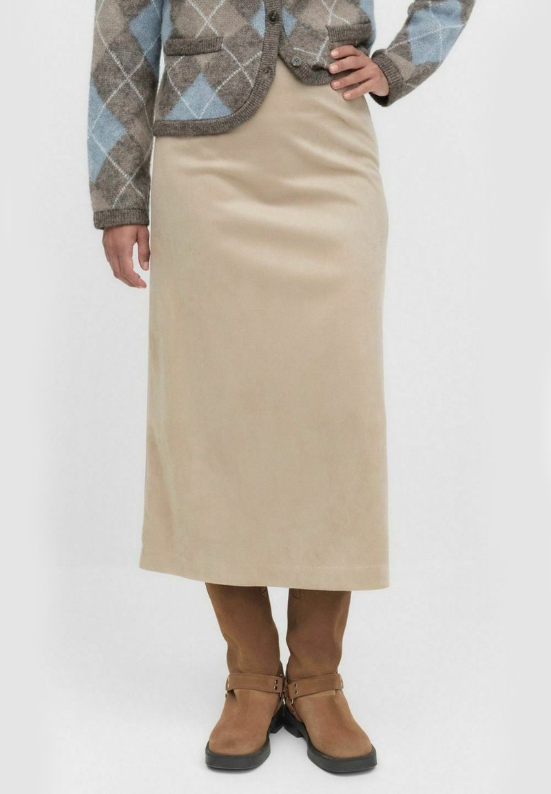 Kvinna i en beige midiklänning, bruna mockastövlar och en brun och blå rutmönstrad cardigan med ena handen på höften.