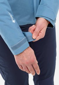 Veste imperméable bleu clair avec des poignets ajustables en Velcro, portée par une personne. Un pantalon marine foncé est partiellement visible. Gros plan sur l'ajustement des poignets.