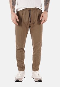 Pantaloni marroni a sigaretta con vita elastica e cordino. Tessuto liscio con un leggero lucido. Abbinati a sneakers sportive bianche.