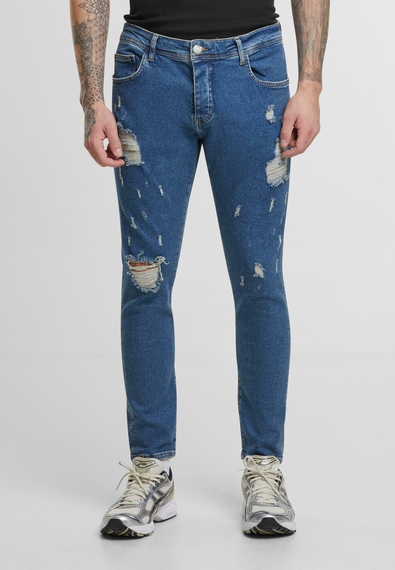 Jean en denim bleu avec une coupe slim, présentant plusieurs accents déchirés et des bords effilochés. Cousu à la machine avec un design classique à cinq poches.