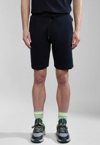 Pantalones cortos de algodón negros con cinturilla ajustable, hasta la rodilla. Usados con zapatillas grises y calcetines verde claro, de pie sobre un fondo gris claro.