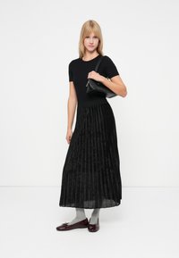 Zwarte geplooide maxi rok met een aansluitend topje, gecombineerd met grijze sokken en glanzende bordeauxrode schoenen. Houdt een kleine zwarte handtas vast.