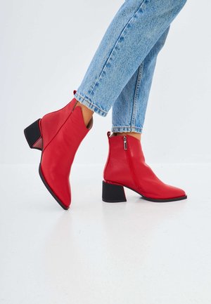 Persona che indossa stivaletti rossi vivaci con tacco a blocco e cerniere, abbinati a jeans di denim azzurro chiaro su uno sfondo bianco semplice.