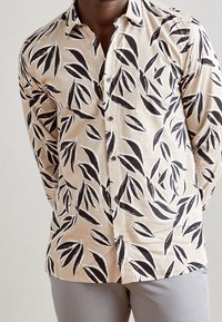 Camisa abotonada de tela beige con un patrón de hojas negras, manga larga y un corte holgado. Detalles en botones oscuros.