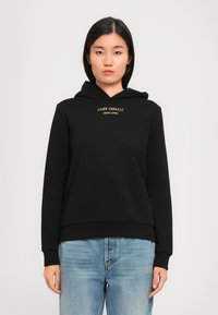 Jeune femme portant un sweat à capuche noir avec le texte "JUST CAVALLI TRUE LOVER", associé à un jean bleu clair, se tenant devant un fond clair.