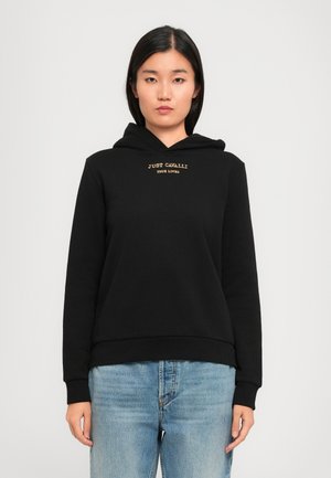Ung kvinne iført svart hettegenser med teksten "JUST CAVALLI TRUE LOVER", kombinert med lyseblå jeans, står foran en ensfarget lys bakgrunn.