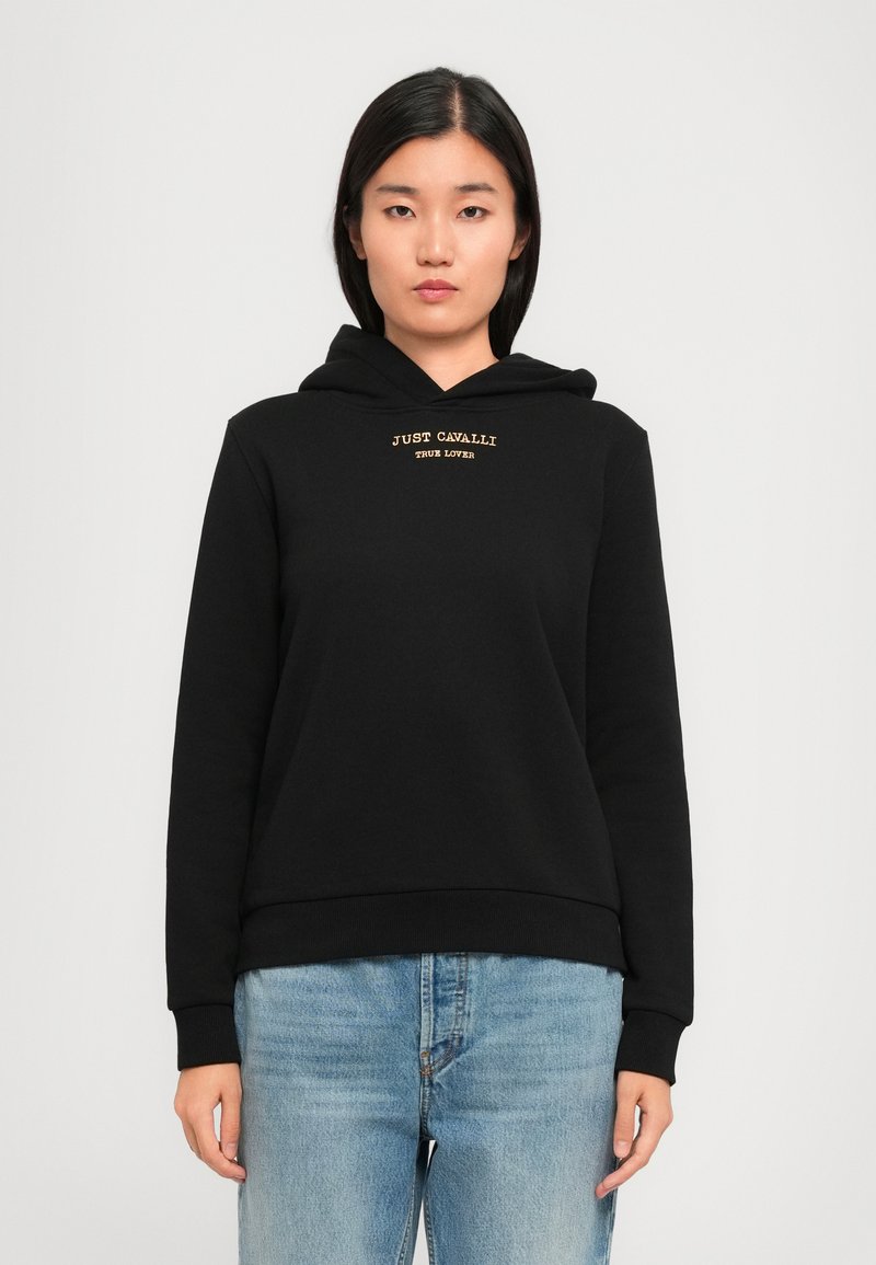 Jeune femme portant un sweat à capuche noir avec le texte "JUST CAVALLI TRUE LOVER", associé à un jean bleu clair, se tenant devant un fond clair.