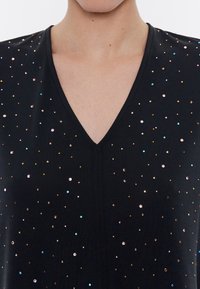 Černý top s výstřihem do V, vyrobený z hladké tkaniny, zdobený barevnými flitry různých velikostí rozptýlenými po látce.