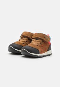 Pantofi pentru copii din suede maro, cu talpă din cauciuc negru, având accente roșii și albastre, o curea cu velcro și un model texturat pe laterale.