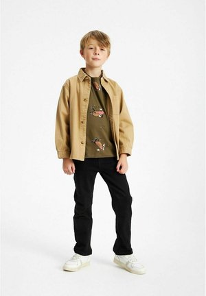 Camicia beige button-up sopra una t-shirt verde con design di cartoni animati, jeans neri e sneakers bianche, in piedi su uno sfondo semplice.