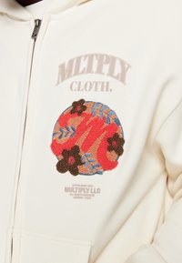 Sudadera con capucha color crema con un diseño bordado texturizado y multicolor que incluye elementos florales y texto de la marca en el frente. Se observa un detalle de cremallera.