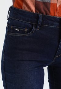 Jeans de mezclilla azul oscuro con un corte ajustado y un ligero desgaste. Presentan un pequeño bolsillo frontal con un logo plateado y detalles de costura estándar.