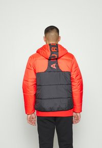 Herrarnas pufferjacka med röd ovandel och svart underdel, med huva och framträdande "AURE"-tryck på baksidan. Slät, kviltad textur.