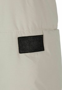 Világos bézs szövet, amelyen egy textúrázott fekete folt látható a márka logójával és szövegével. A varrás részletei láthatóak; az anyag lágy és könnyű érezhető.