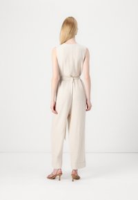 Combinaison en lin beige avec un design sans manches, taille ceinturée, jambe large et poignets retroussés, assortie à des talons à lanières. Fond uni.