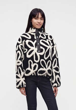 Femme portant un pull polaire noir et blanc avec de grands motifs floraux abstraits et des boutons-pression sur la patte de boutonnage avant.