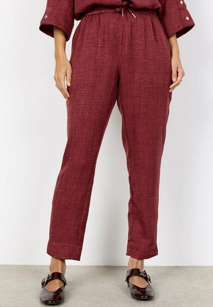 Vrouw draagt een gestructureerde bordeauxrode broek met een trekkoord in de taille en donkere glanzende Mary Jane schoenen, staand op een grijze vloer.