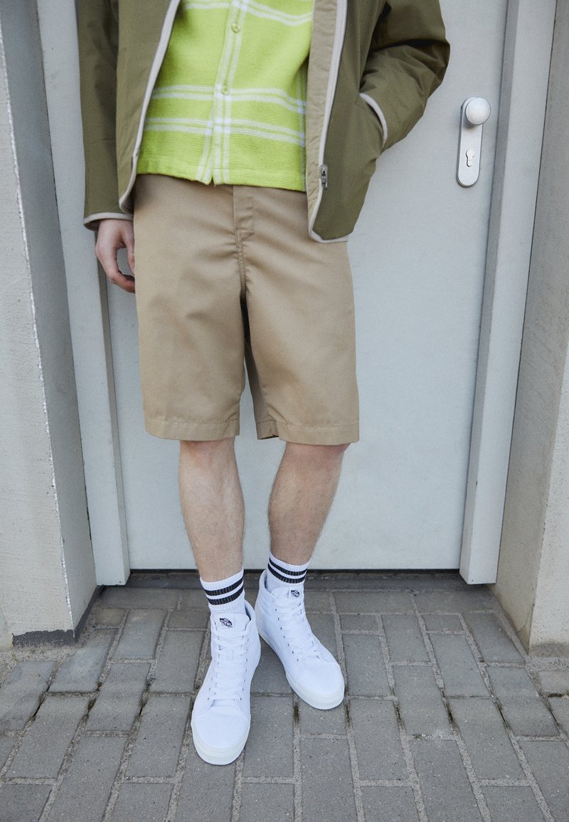 Personne debout portant un short beige, des baskets blanches montantes et des chaussettes blanches à rayures noires, vêtue d'une veste verte et d'une chemise, devant une porte.