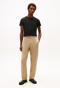 Črna bombažna majica z majhnim logotipom, kombinirana z bež hlačami z ravnim krojem in sprednjimi žepi, nošena s črnimi čevlji.