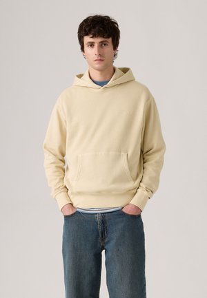 Crèmekleurige hoodie met een voorzak, zachte textuur en geribbelde manchetten. Draag het over een blauwe shirt en gecombineerd met een donkere spijkerbroek.