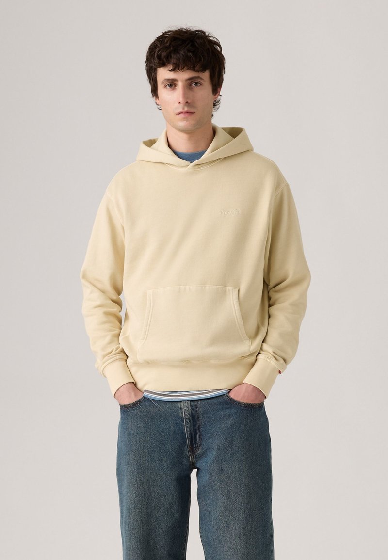 Crèmekleurige hoodie met een voorzak, zachte textuur en geribbelde manchetten. Draag het over een blauwe shirt en gecombineerd met een donkere spijkerbroek.