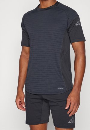 Homme portant un t-shirt de sport noir à manches courtes et un short avec des logos Adidas discrets, debout devant un fond clair uni.