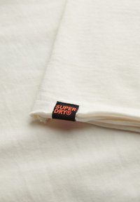 Tissu en coton blanc plié, avec une étiquette noire portant "SUPER DRY" en lettres orange près du bord. Texture lisse et design léger.