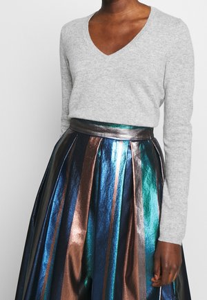 Grauer V-Ausschnitt-Pullover aus weichem Strick, kombiniert mit einem metallischen, plissierten Rock mit blauen, grünen und bronzefarbenen Streifen und einem glänzenden Bund.