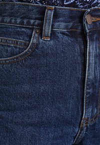 Tmavě modré džíny z denimu s oranžovým prošíváním, mají přední kapsu a kovové zapínání na knoflík v pase. Rovnoměrná struktura.