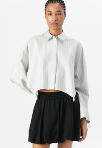 Chemise à manches longues légèrement raccourcie en gris clair avec des rayures blanches verticales, associée à une jupe noire à taille élastique, en tissu doux et fluide.