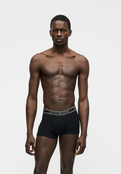 Izmos férfi rövid hajjal, fekete Emporio Armani boxeralsót viselve, egy egyszínű világosszürke háttér előtt áll, előre nézve.