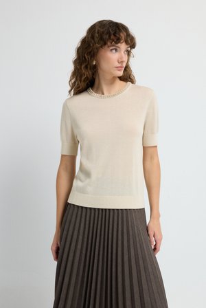 Beige kortinmouwen trui met een geribbelde halslijn en manchetten, gecombineerd met een geplooide bruine rok. Zachte textuur, aansluitend ontwerp en effen kleur.