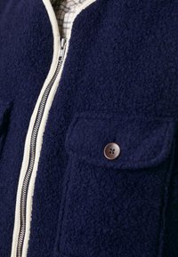 Jūras spēku zila vilnas maisījuma jaka ar maigu tekstūru, ar rāvējslēdzēja aizdari, krēmkrāsas apdari un priekšējo kabatu ar brūnas pogas detaļu.