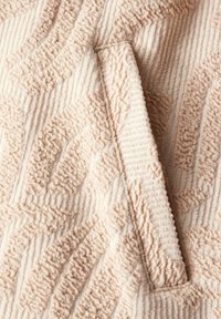 Tela texturizada beige que presenta un patrón en relieve y un diseño de bolsillo prominente. La superficie tiene estrías distintivas y suavidad.