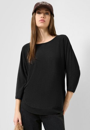 Frau mit brauner Mütze, schwarzem locker sitzendem Langarmshirt und schwarzer Hose steht vor einem schlichten hellen Hintergrund.