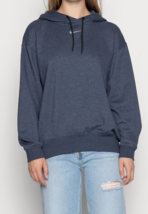 Persoon draagt een donkerblauwe Nike hoodie met zwarte trekkoorden en lichtblauwe gescheurde jeans, staand tegen een effen achtergrond.
