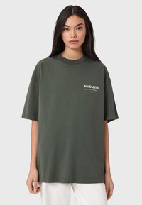 Ylimitoitettu oliivinvihreä puuvillainen T-paita, jossa on valkoinen logo vasemmalla rinnassa, pyöreä pääntie ja lyhyet hihat. Sileä koostumus, rento istuvuus.