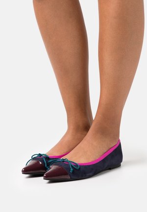 Flats en daim bleu marine avec bouts pointus brillants bordeaux, accents de nœud bleu sarcelle et bordure rose vif portés pieds nus sur fond blanc.