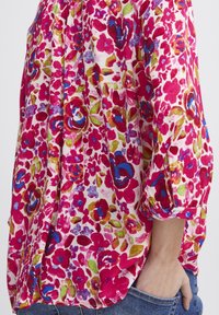 Blommig skjorta i rosa, röd och grön nyanser; lättviktig tyg med avslappnad passform; har uppkavlade ärmar och en knapp detalj på manschetten.