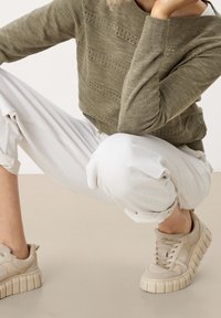 Pull en maille vert olive avec des détails texturés horizontaux, associé à un pantalon blanc ample et des baskets beiges à semelle épaisse.