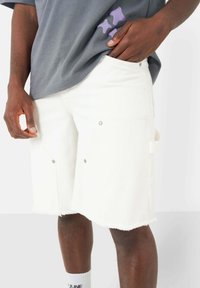 Shorts in denim bianco con orlo strappato, tasche laterali e dettagli in metallo argentato. Abbinati a una t-shirt grigia con una grafica viola.