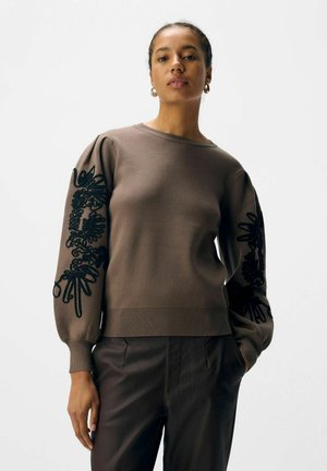 Maglione marrone con maniche a sbuffo, decorato con ricami floreali neri sulle maniche. Collo e orlo a costine. Indossato con pantaloni scuri.