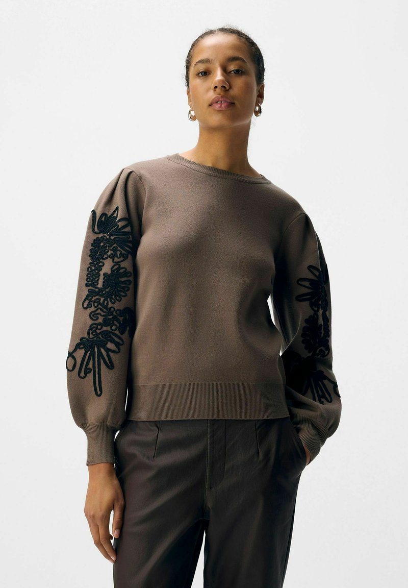Pull marron avec des manches bouffantes, orné de broderies florales noires sur les manches. Col et ourlet côtelés. Porté avec un pantalon sombre.