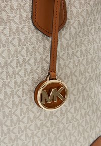 Sangle en cuir marron avec breloque logo MK dorée Michael Kors sur sac en tissu beige à motif répétant les initiales MK.