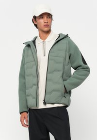 Veste en polaire verte avec texture matelassée et capuche, présentant une fermeture éclair sur le devant et des accents noirs, portée par-dessus un polo beige et un pantalon foncé.